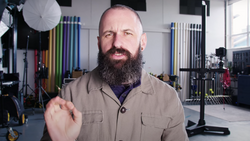 Bearded man in tan shirt gesturing while speaking in a photography studio with colorful light stands and softbox visible in background.
