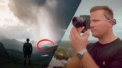 Photographer holding a camera on a mountainside as another figure stands in a distant valley below dramatic cloudy skies.