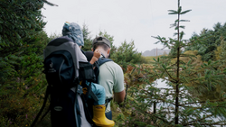 Backpacker wearing large hiking pack walks along forested trail beside calm water.