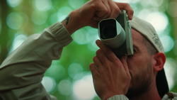 Photographer holding a rangefinder camera to their eye against a soft, blurred green foliage background.