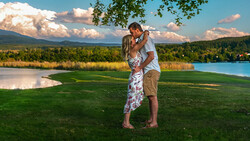 Couple kissing in a grassy field with mountains and lake in the background.