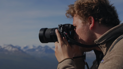 Photographer with curly reddish-brown hair holds a mirrorless camera with telephoto lens to his face, composing a shot of snow-capped mountains under clear blue sky.