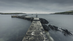 Long stone pier extending into calm gray water with rocky outcrops and distant coastline under overcast sky.