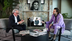 Two men seated across from each other in a studio setting with framed photographs and memorabilia on a table between them.