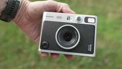 Hand holding a Fujifilm Instax hybrid instant camera with a black textured body and silver trim against a blurred green background.