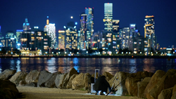 Nighttime cityscape with illuminated skyscrapers reflected in water, foreground shows silhouetted figure fishing among rocky breakwater.