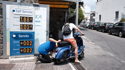 Person bending over a blue Vespa scooter parked in front of a gas station price sign on an Italian street.