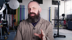 Bearded man in tan work shirt gesturing while speaking in a professional studio setting.