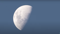Waning crescent moon against a clear blue sky, surface details and craters visible in sharp focus.