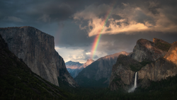 Rainbow arcing over a dramatic mountain valley at sunset with granite cliffs and a waterfall.
