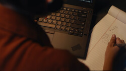 Person in rust-colored sleeve pointing at handwritten notes beside a laptop keyboard during evening work session.