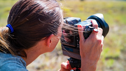 Photographer holding a video camera to their eye while operating it outdoors in daylight.