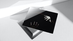 Black card with white hands reaching toward each other, positioned on white geometric surfaces with dramatic diagonal lighting.