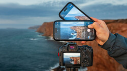 Hand holding smartphone and DSLR camera both displaying the same coastal cliff landscape with ocean.
