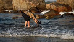 Photographer in striped shirt bending forward in shallow ocean water while holding a camera on a rocky coastline.
