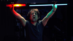 Man holding illuminated neon tubes overhead with arms raised in celebration against dark background.