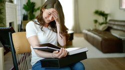 Woman sitting indoors holding stacked books with hand to forehead, appearing stressed or overwhelmed.
