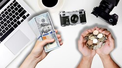 Hands holding US currency and coins next to vintage cameras and laptop on white surface.