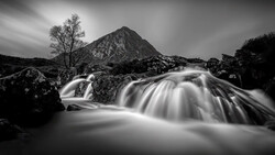 Long-exposure photograph of a rushing mountain stream with a bare tree and rocky peak in the background.