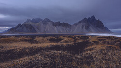 Dramatic mountain range with dark peaks rising above golden tundra landscape under moody twilight sky.