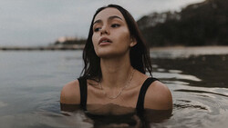 Woman in black swimsuit partially submerged in calm water, gazing upward with a contemplative expression.