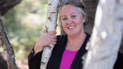 Woman smiling at camera while holding a birch tree trunk in natural outdoor lighting.
