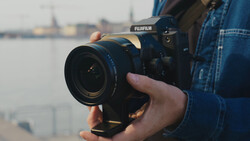 Photographer holding a Fujifilm mirrorless camera with attached lens, positioned toward a waterfront cityscape.