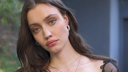 Woman with long dark hair tilting her head slightly, wearing pearl earrings and a delicate chain necklace.