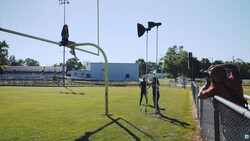 Two people standing on a sunny baseball field with tall light poles and a blue building visible in the background.