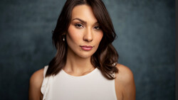 Woman with dark wavy hair wearing white sleeveless top against dark blue-gray backdrop.