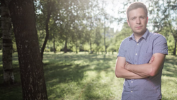 Man in light purple shirt standing with crossed arms in a park with tall trees and dappled sunlight.