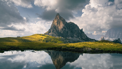 Dramatic mountain peak reflected in still water with golden meadow and cloudy sky.