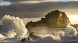 Large ocean waves crash against a rocky sea stack during golden hour with dramatic storm clouds overhead.