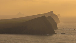 Dramatic coastal cliffs shrouded in golden mist, their steep faces rising from calm waters in atmospheric conditions.