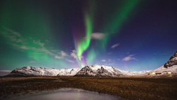 Northern lights dancing in green and purple bands above snow-capped mountains and a frozen lake.