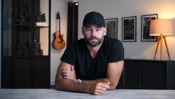 Man in black cap and shirt leaning against table in studio with warm lighting and acoustic guitar visible in background.