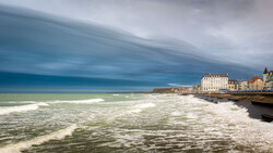 Dramatic coastal town with white-capped waves and moody sky reflected in water.