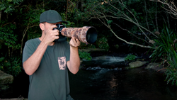 Photographer with telephoto lens in forest setting near water.