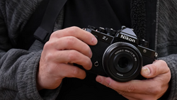 Hands holding a black Nikon film SLR camera with a standard lens against a dark textured jacket.