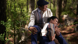 Adult and child in embroidered traditional clothing crouching together in a forested setting.