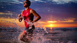 Male athlete running through ocean water at sunset wearing red swimwear and sunglasses.
