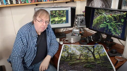 Photographer reviewing a forest landscape print at his desk workstation with dual monitors.