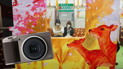 Ricoh GR camera displayed in front of a colorful mural featuring red deer and autumn foliage.