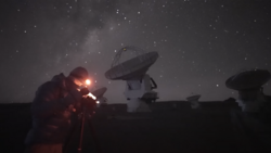 Radio telescope dishes silhouetted against a starry night sky with an explosion or bright light source illuminating one telescope in orange and red.