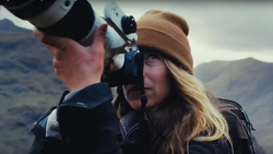 Photographer with long blonde hair wearing a tan beanie looks through a camera viewfinder while holding binoculars in a mountainous landscape.