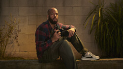 Photographer sitting against concrete wall holding camera, wearing plaid shirt and sneakers.