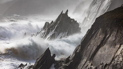 Dramatic coastal rock formations surrounded by crashing waves and sea spray in monochromatic tones.