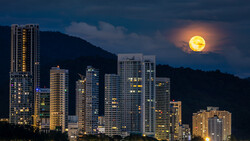 Illuminated high-rise buildings silhouetted against a full moon rising over mountains at dusk.