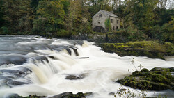 Flowing waterfall cascading over rocks with a stone mill building nestled among trees on the riverbank.