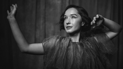 Woman with arms raised looking upward in a black and white portrait against a draped backdrop.